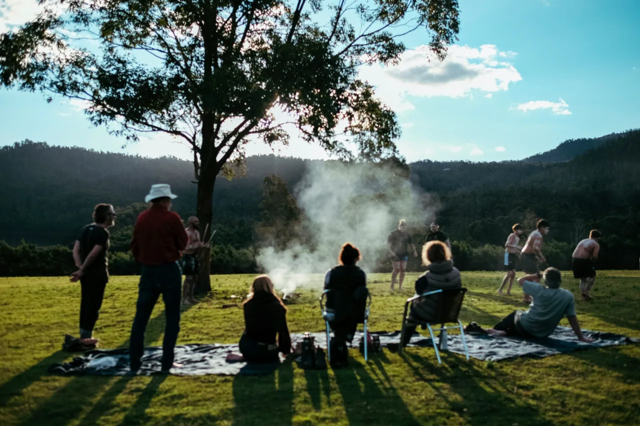 A group seated around a fire in an open paddock, escarpment behind.