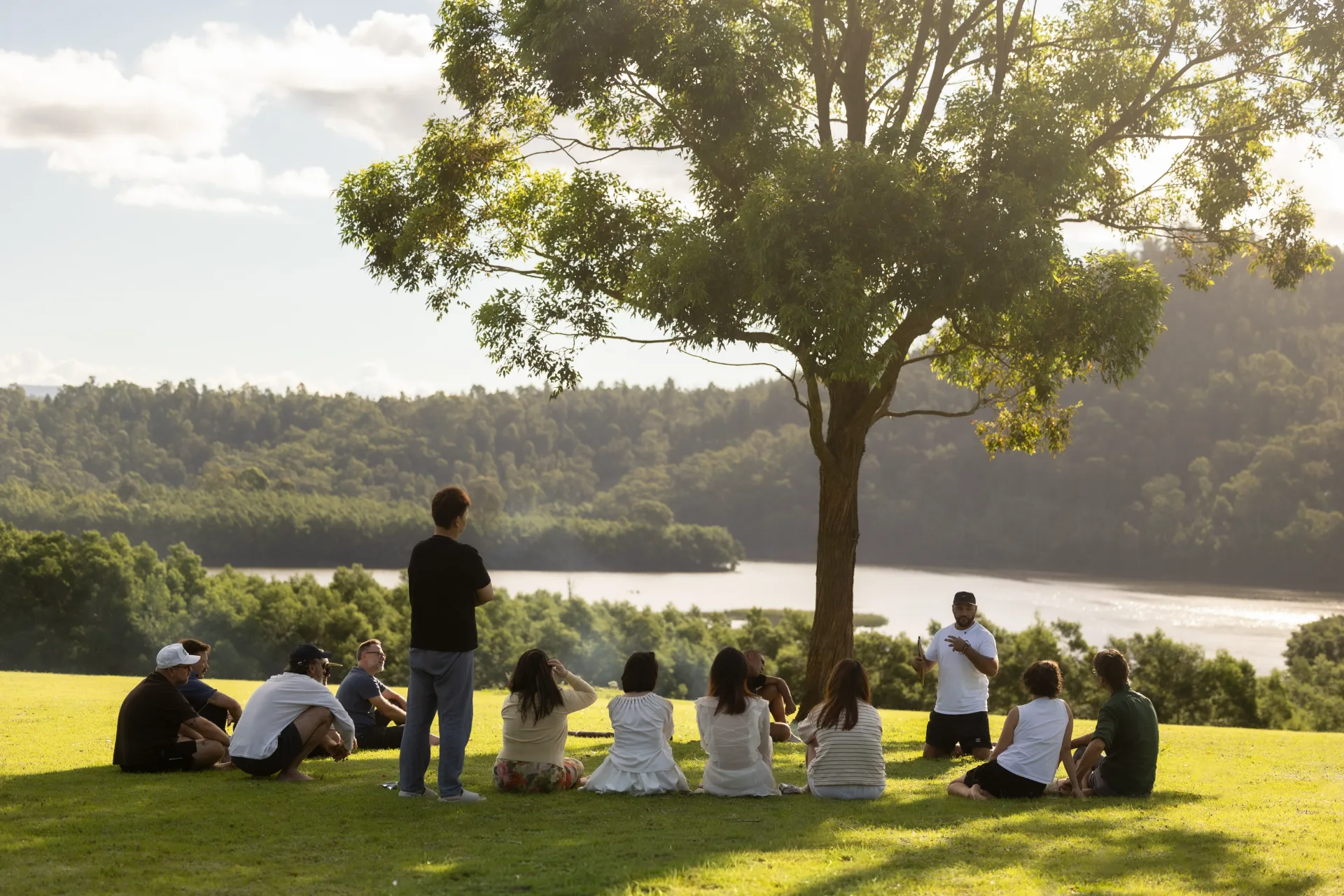 A small group gathered on the grass under a tree above the water.