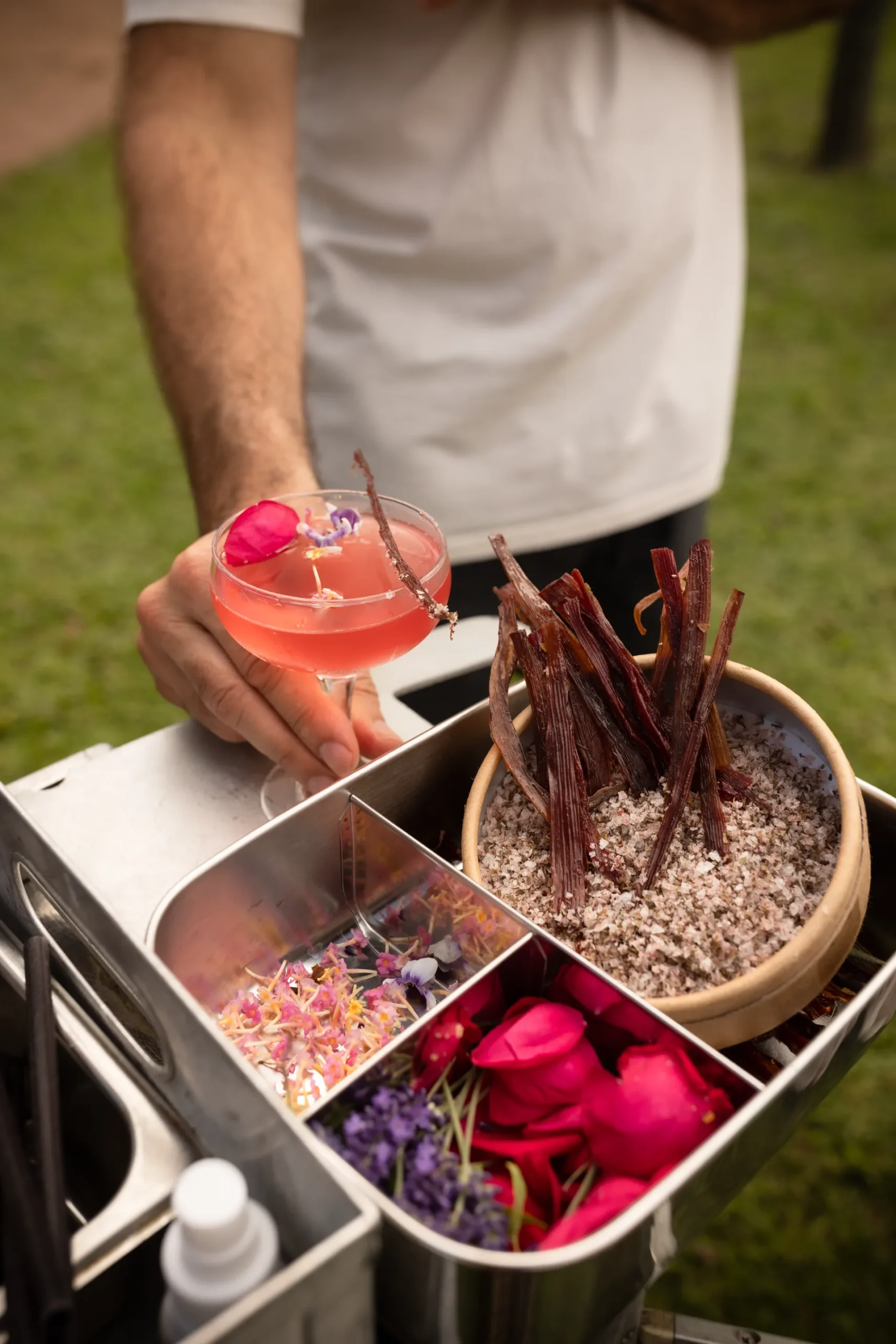 A coupe of pink botanical aperitif held beside a tray of native flowers, rose petals and bark.