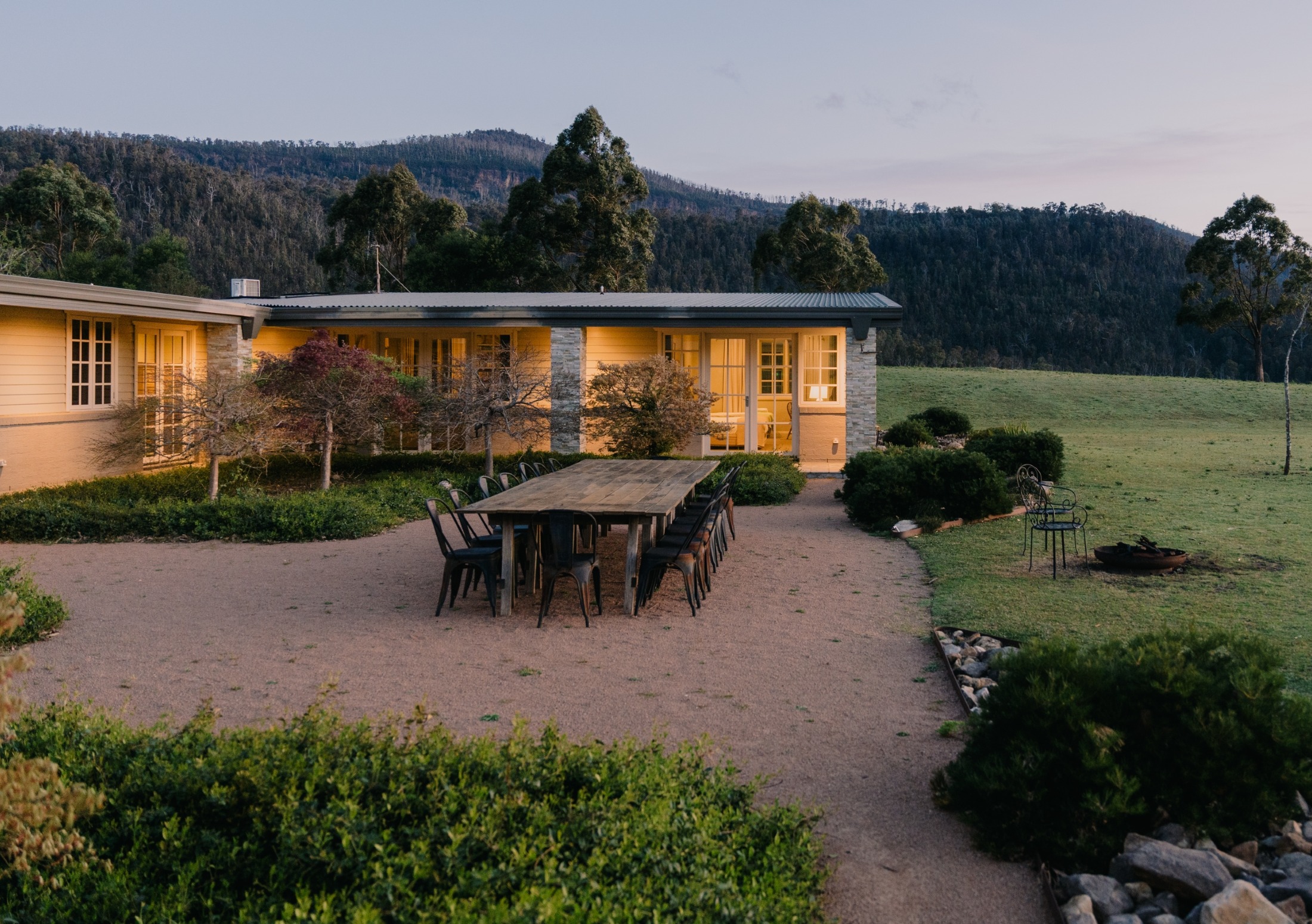 The lodge at dusk with its windows lit, a long table set outside on the gravel, and forested hills behind.