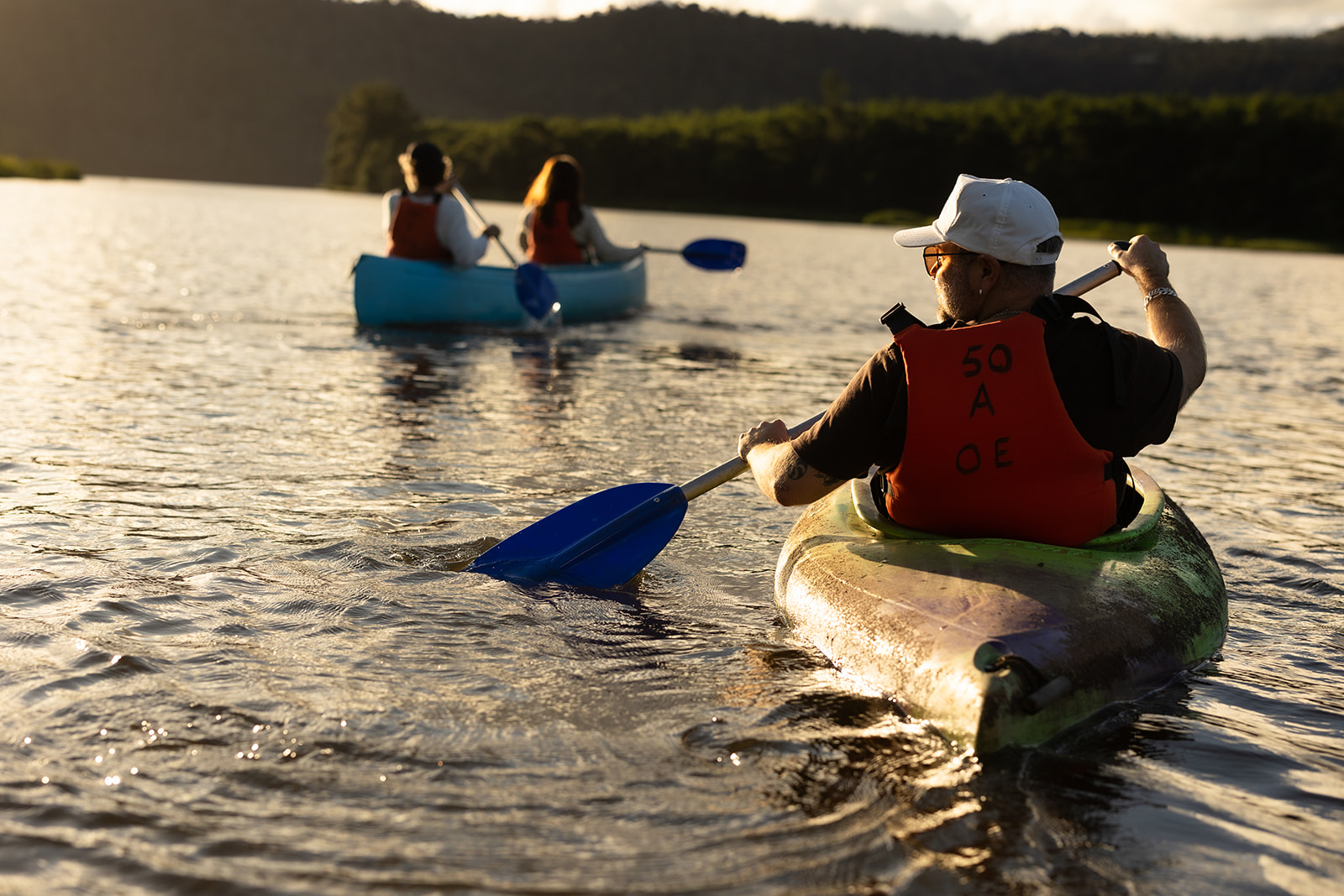 Three people paddling on flat water in late afternoon light, the far bank wooded behind them.